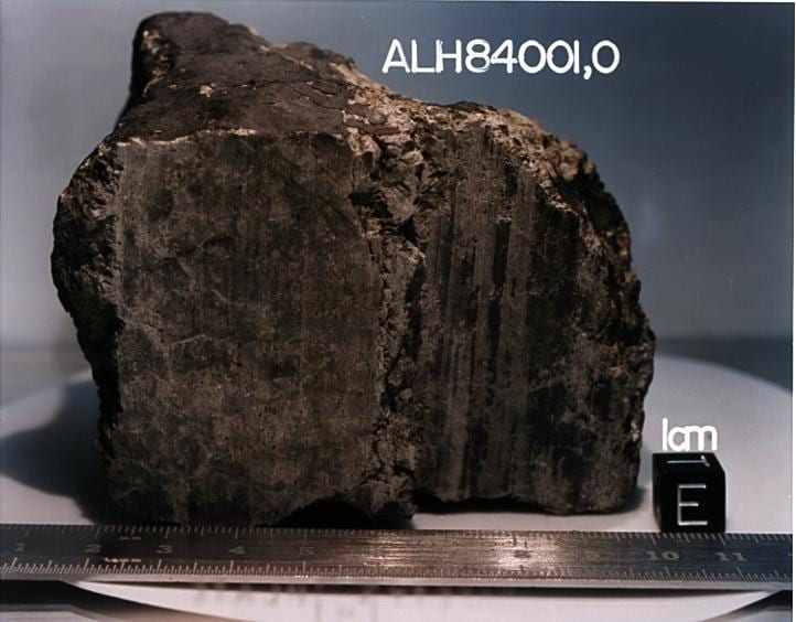 A dark, fist-sized meteorite resting on a white surface beside a scale cube, its sawn face showing a fine black fusion crust.