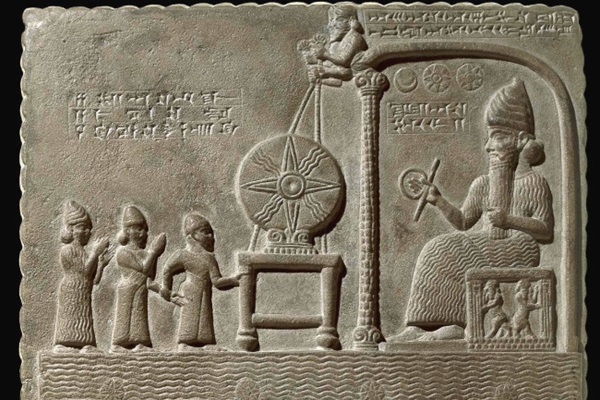 Carved stone relief: a bearded god enthroned under a canopy holding a rod and ring, a large rayed disc on an altar before him, three smaller figures approaching from the left.