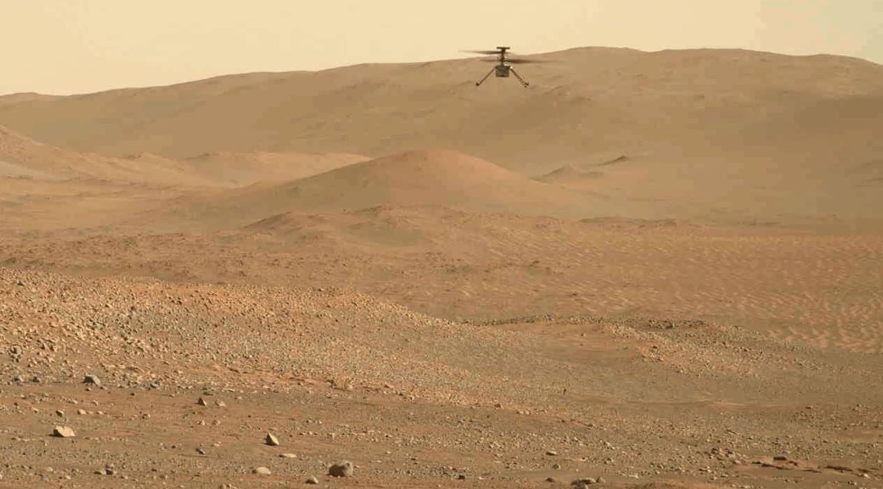 A wide Martian landscape of low dunes and scattered rock, with a small four-rotor helicopter airborne above the ridge line.