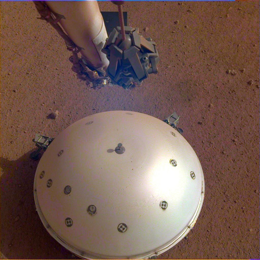 A white domed shield sitting on reddish Martian soil, with the lander's robotic arm and grapple hanging in frame above it.