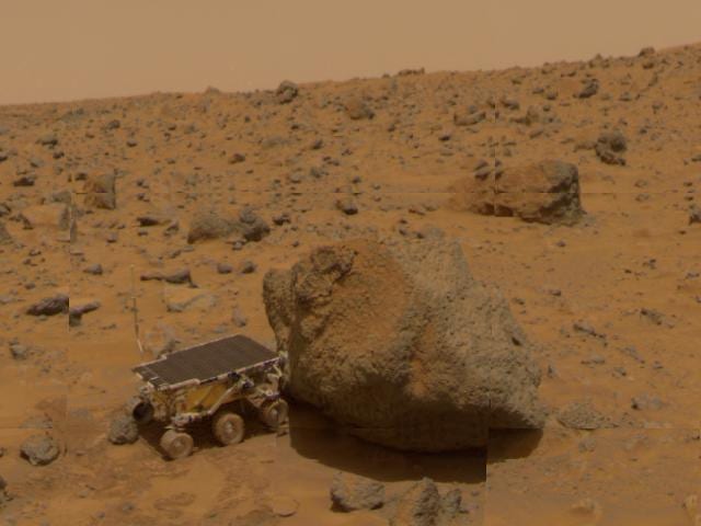 A small six-wheeled rover parked against a large rounded boulder on a rock-strewn orange plain.