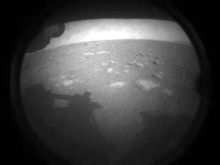A low, wide-angle black and white view of flat Martian ground, seen through a dust-speckled lens cover.
